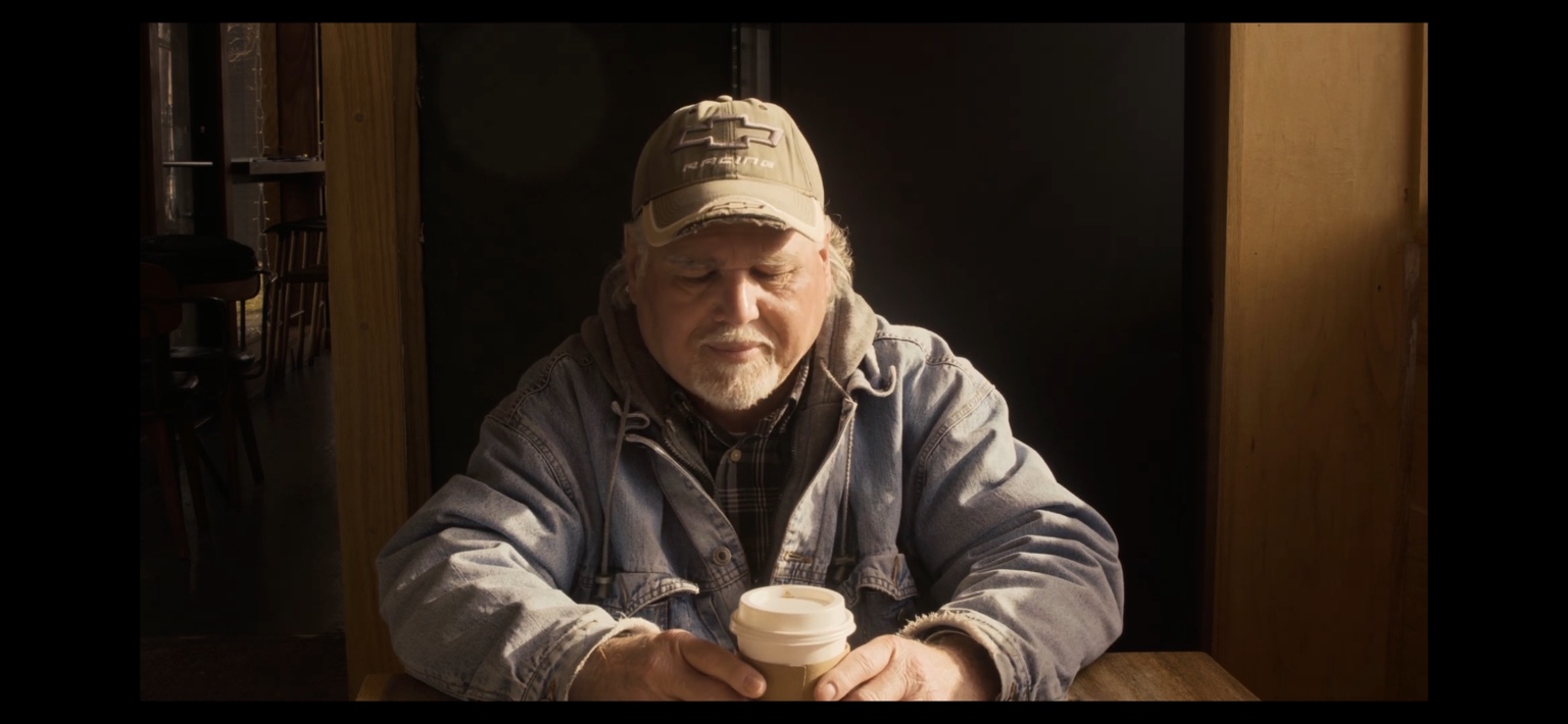 A man sits with a coffee in a warmly lit frame from The Coffee Shop.